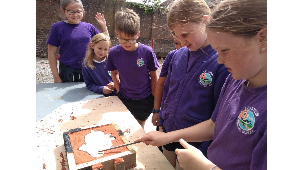 A row of children inspecting a metal cast still in the sand mould
