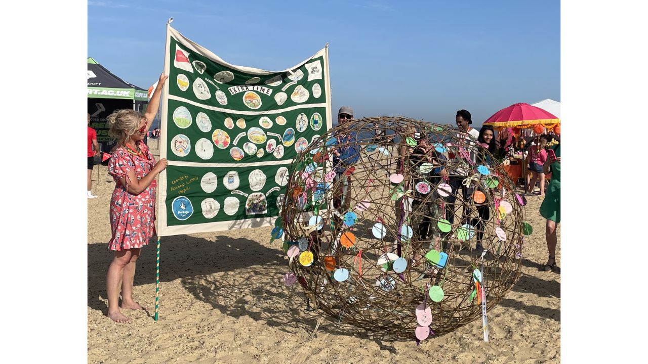 Beach scene with adults holding large green and white banner and decorated willow ball in foreground with people standing round