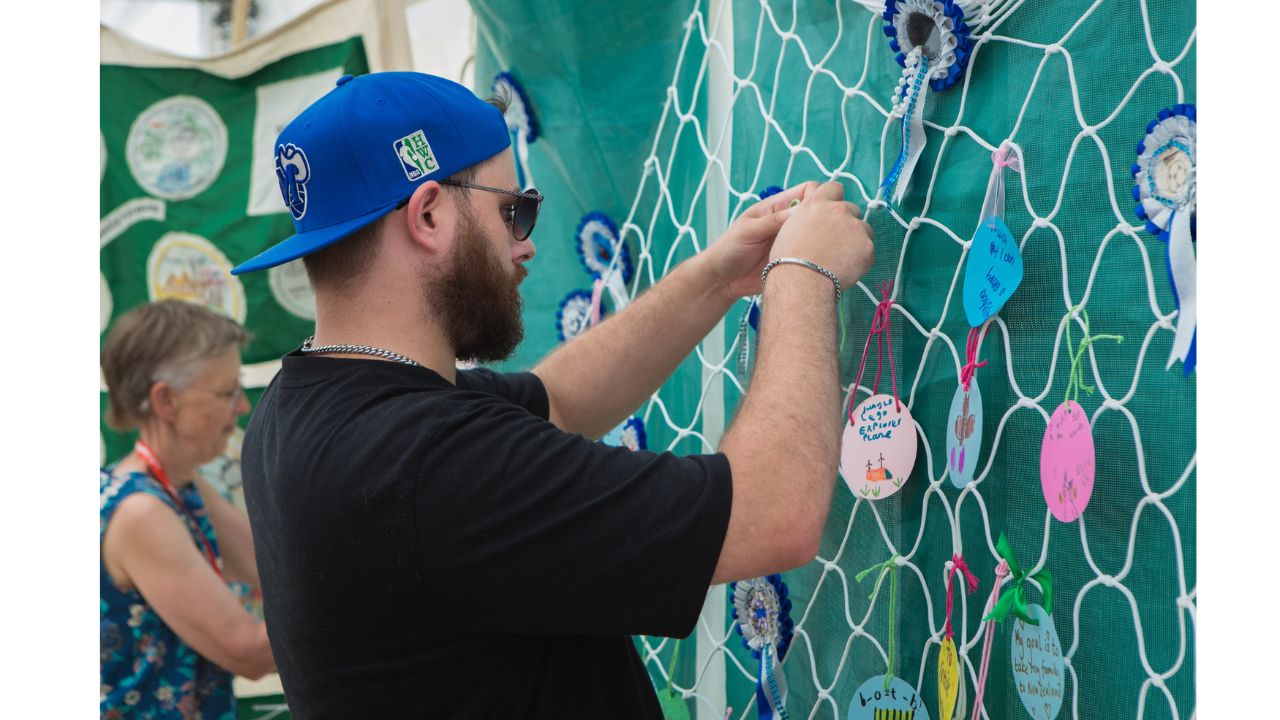 A bearded man wearing sunglasses and a blue baseball cap backwards ties something to a large piece of white football net