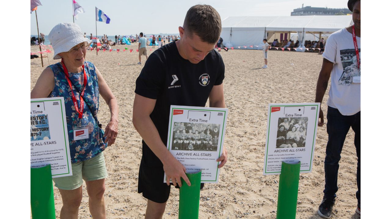 Adults look on as a man in a sports top and shorts places a green tube in the sand. Three tubes each with a board celebrating an Archive All-Star