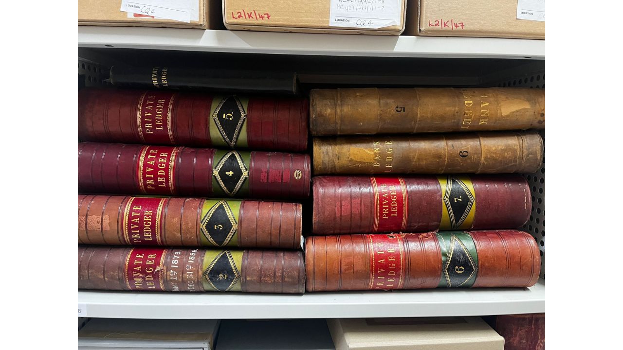 9 old leather bound private ledgers lying horizontally on a shelf with cardboard boxes on shelf abover