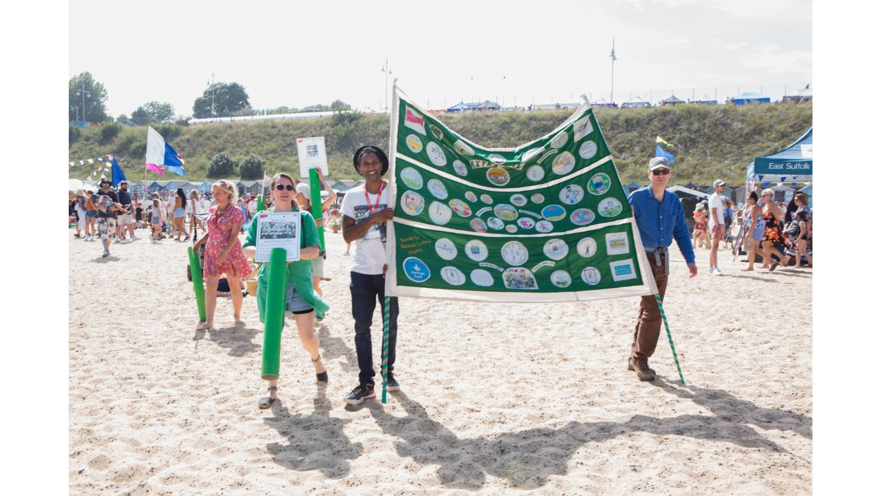 Adults walking across beach casting shadows in strong sunlight and holding large green and white banner