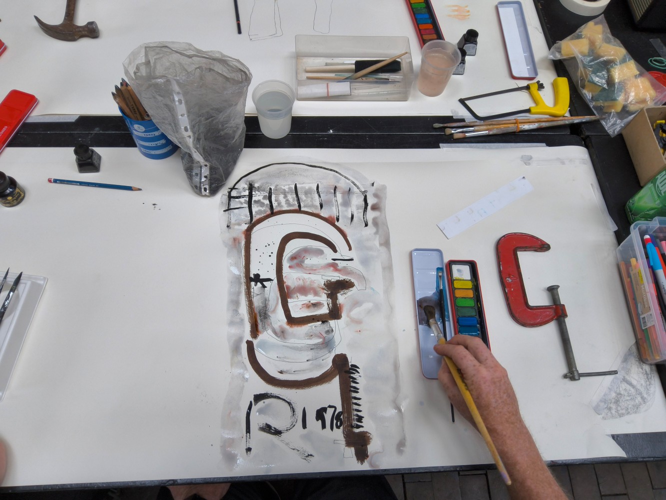 A red metal clamp, a paint box and a painting of the tool, together with other art materials on a white background