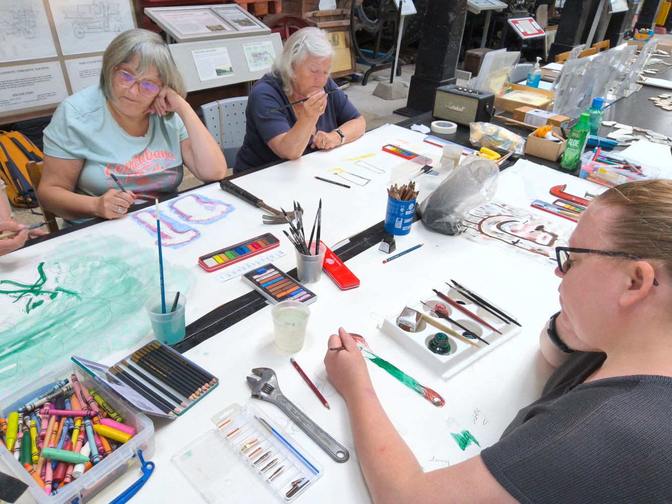 Adults sit either side of a long table covered in white paper and assorted art materials, each drawing a tool
