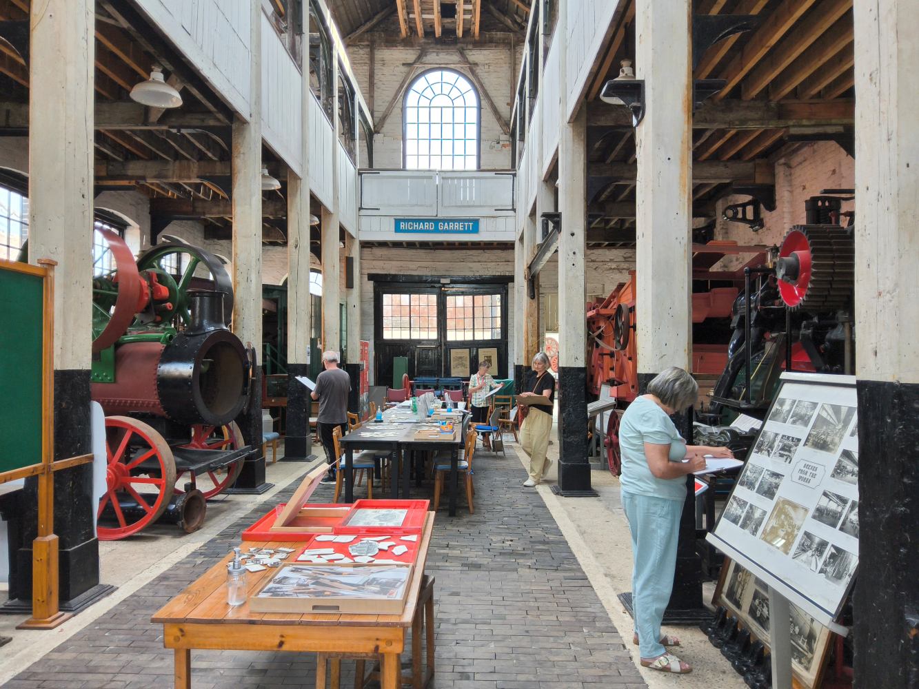 A long view of an old engineering works, with steam engines either side, tables down the middle and people standing and sketching items