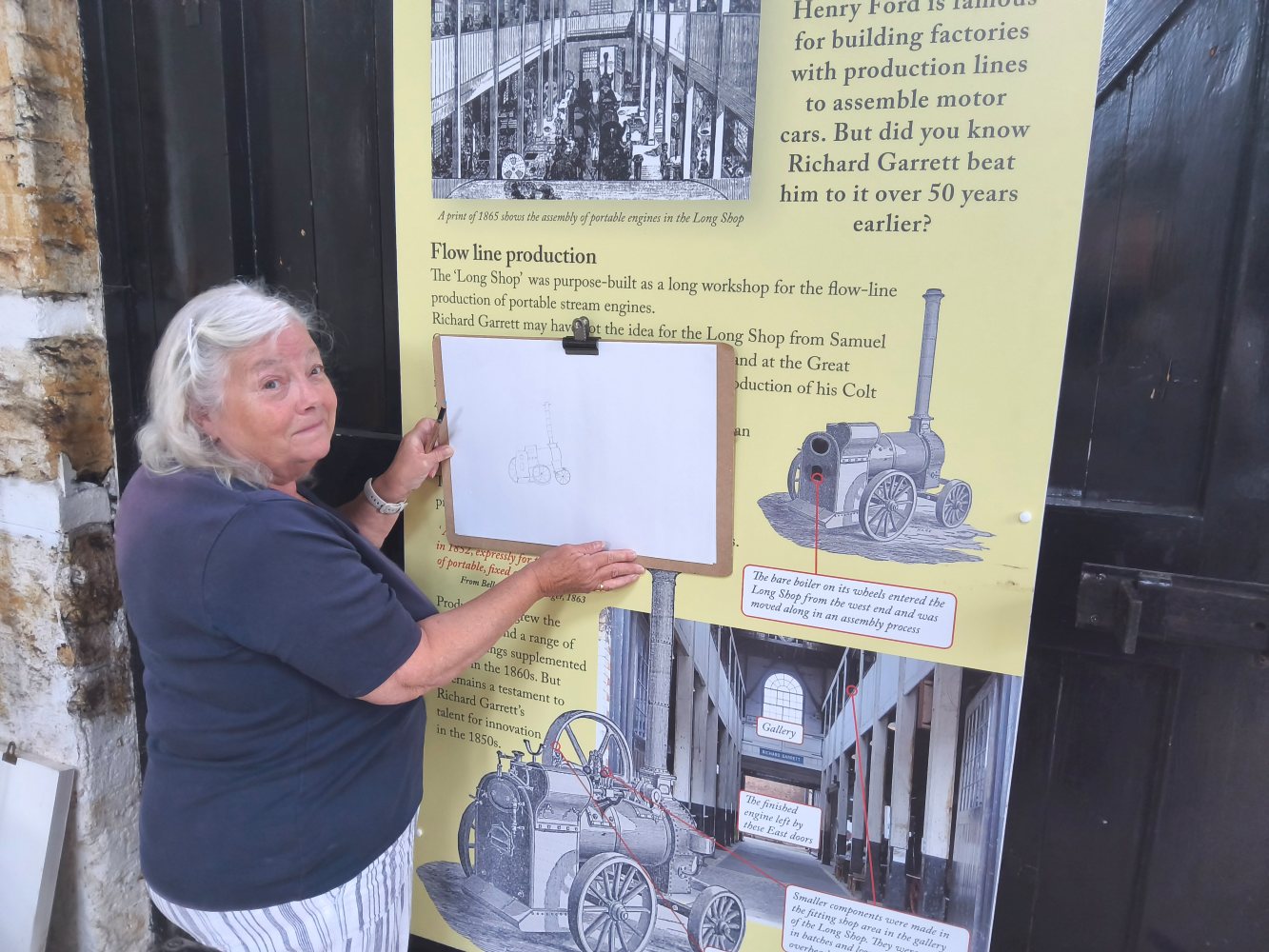 A woman holds up her drawing to compare it with an image of an old steam engine on an information board about flow line production