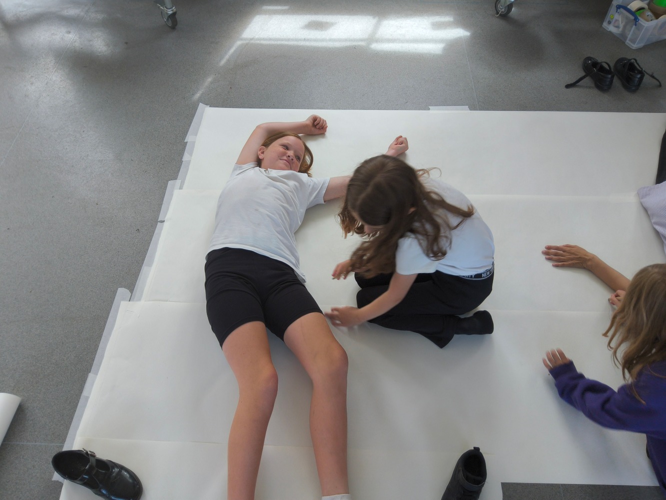 A child dressed in white t-shirt and black shorts lies stretched out on a large piece of white paper whilst another child draws round her