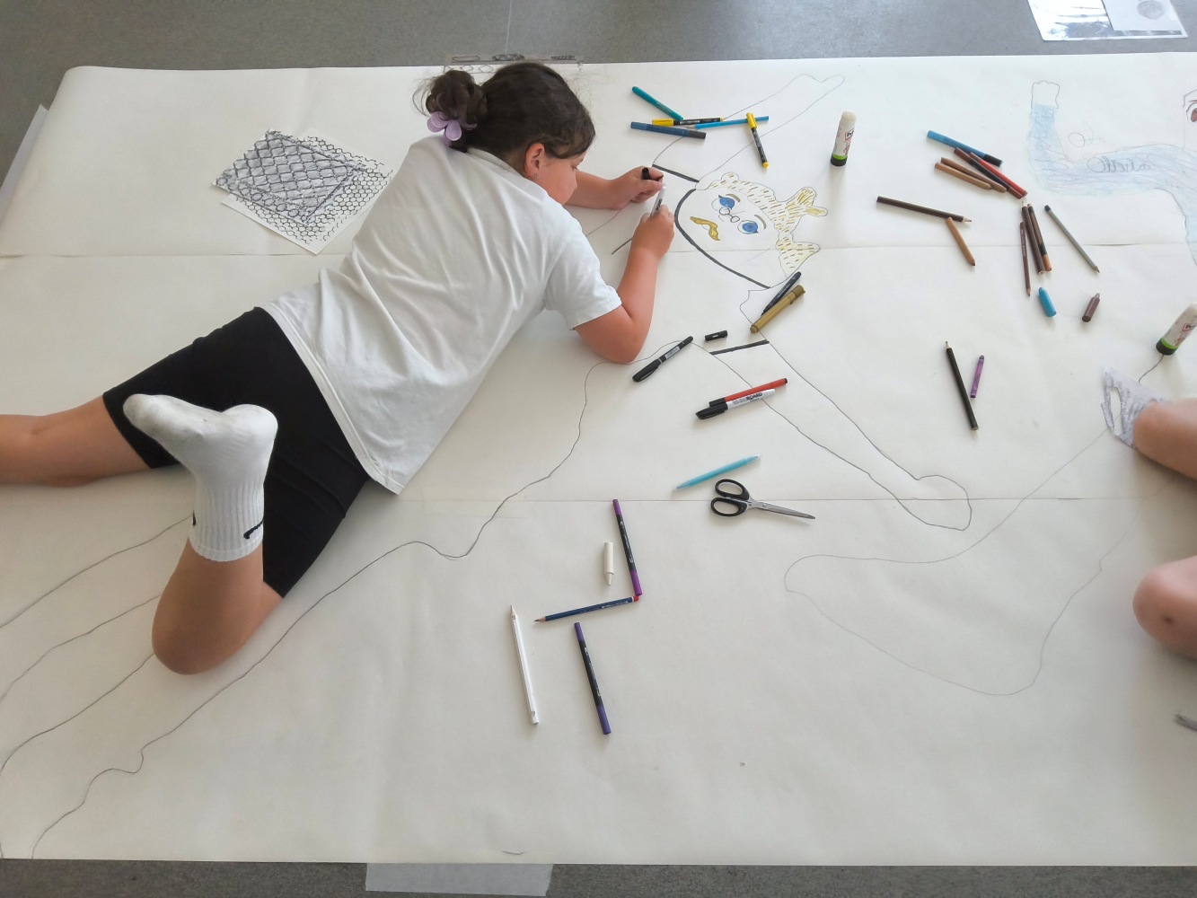 A child lies on her stomach on the life size drawing of herself, with a variety of pens around, as she adds detail to the drawing