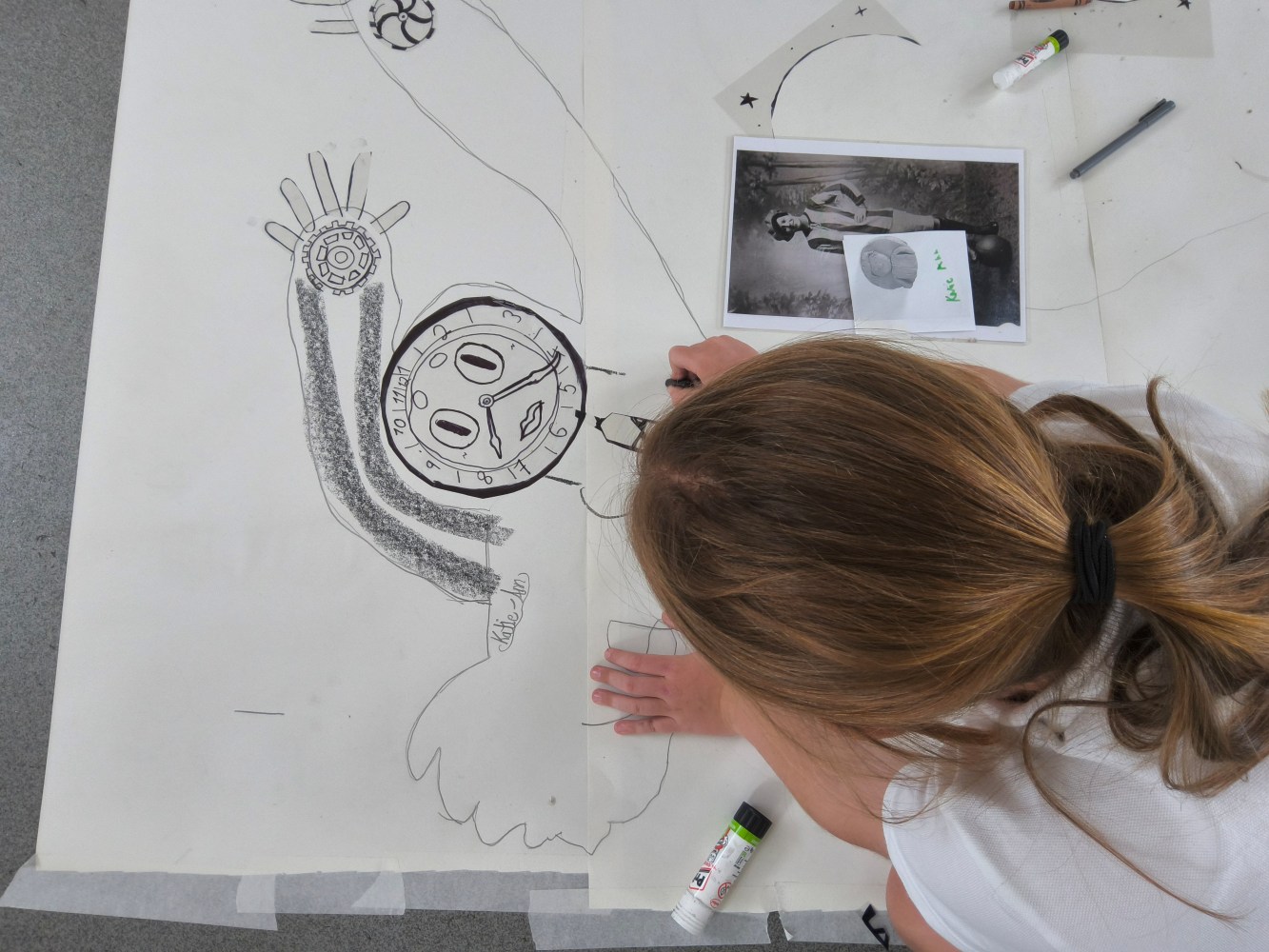 Overhead view of child drawing figure with clock face, arms raised and photo of munitionette beside