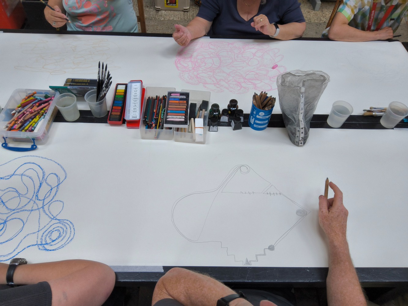 Long strips of white paper laid out on a table with drawing and painting materials down the middle. A series of hands and arms suggest there are people sat either side of the table