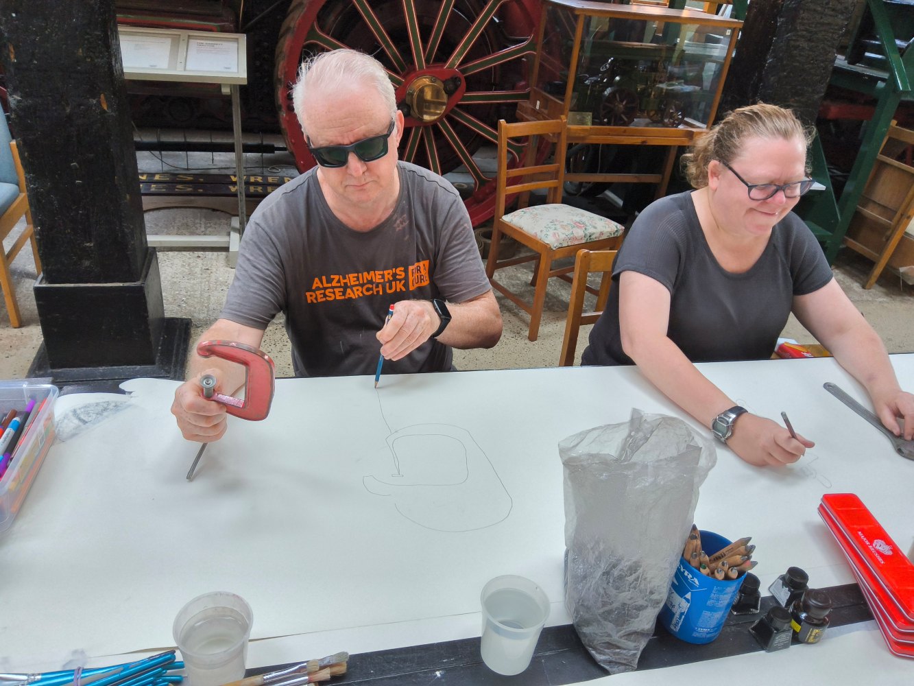 Two adults sit side by side in front of a long piece of drawing paper. Each holds a tool in their hand, the man holds a red clamp and the woman a spanner and they are drawing the objects. Also in front of them are beakers of water, pencils and red paint tins