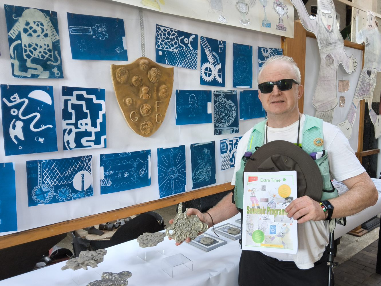 A man in dark glasses and white T shirt stands in front of a display of cyanotypes and a large plaster shield. He holds a publication in one hand and a pewter model in the other
