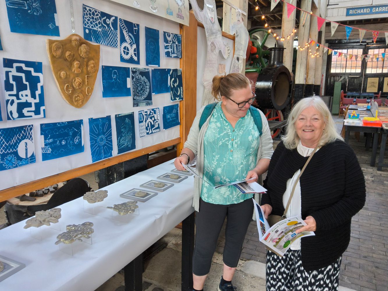 Two women looking at the Souvenir Programme standing in front of a display of cyanotype pictures, a plaster shield and cast pewter models