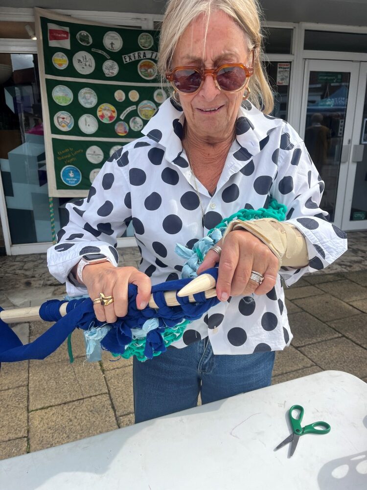 A woman wearing a spotted shirt and sunglasses knits with blue wall on large wooden needles. In the background is a large football banner with embroidered pictures