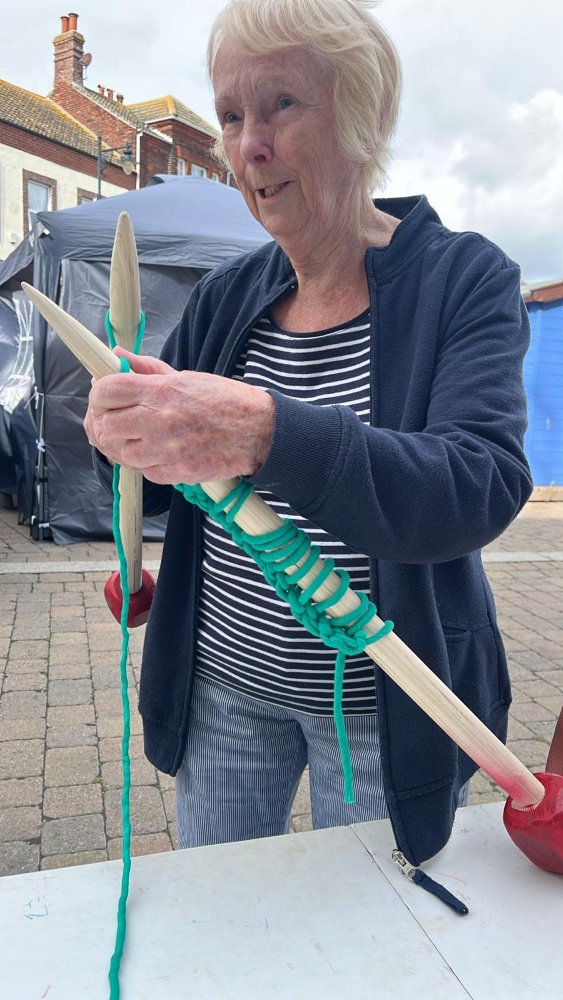 A woman holds a huge pair of wooden knitting needles with a row of green stitches cast on