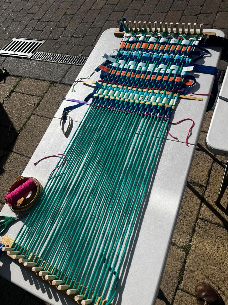 A table top loom, with green wool with ribbons woven through
