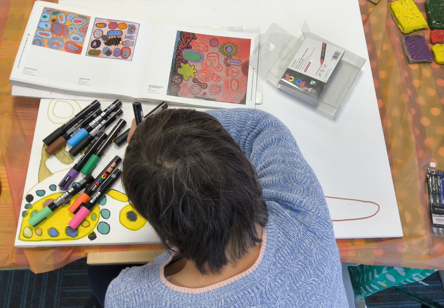 A person leans over a white canvas on which are scattered a variety of paint pens, a empty box and a book opened at a double page spread of work by the Japanese artist Yayoi Kusama