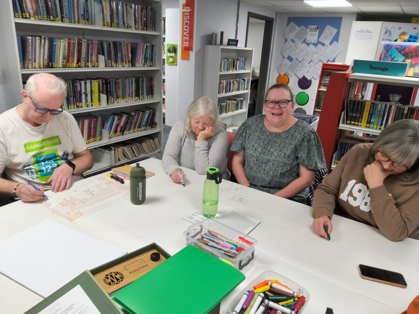 A group of adults sit around a table in a library, with pens and paper in front of them
