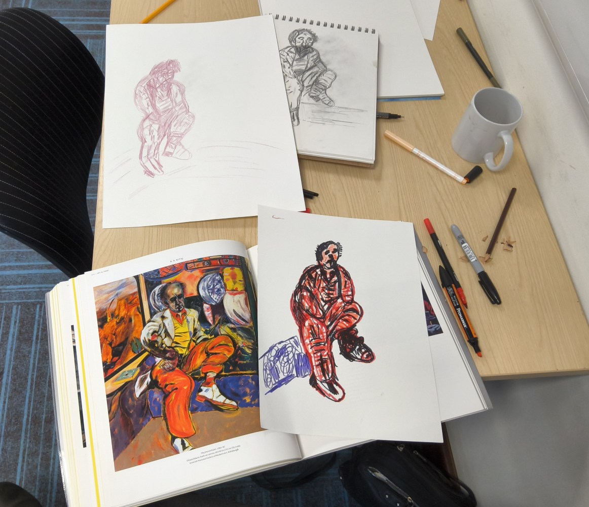 There is a spiral bound sketch book with a graphite picture of a figure, overlaid with a similar sketch in red ink. Beneath these pictures is a book opened at an illustration R B Kitaj and the Brave artist's interpretation of the image. The table is scattered with pens and an empty white coffee mug