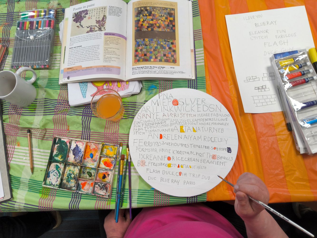 Brightly coloured orange and green plaid table cloths are covered with an art book about Klee, pens, paints and a palate. Centre is a circular canvas on which the artist is creating work inspired by letter shape and colour