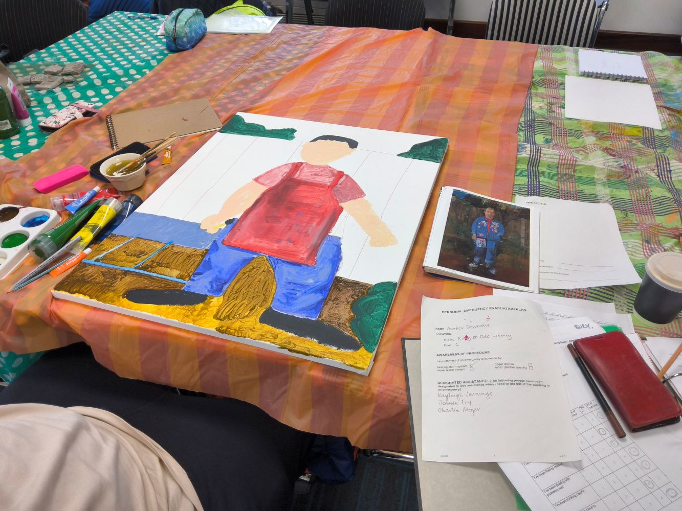 A table covered with multi-coloured cloths with a large canvas showing an unfinished painting, with books and papers alongside