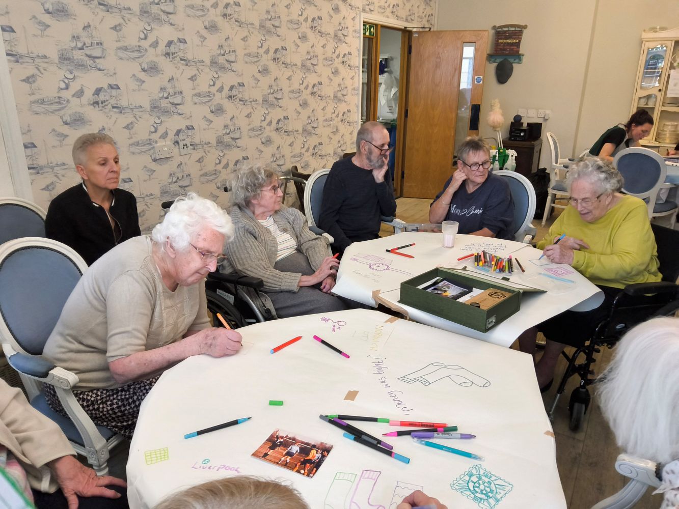 A group of adults gathered round two circular tables, each covered with lining paper. A woman in the foreground leans over the table, writing something whilst another lady in a mustard coloured top draws an image as other adults look on