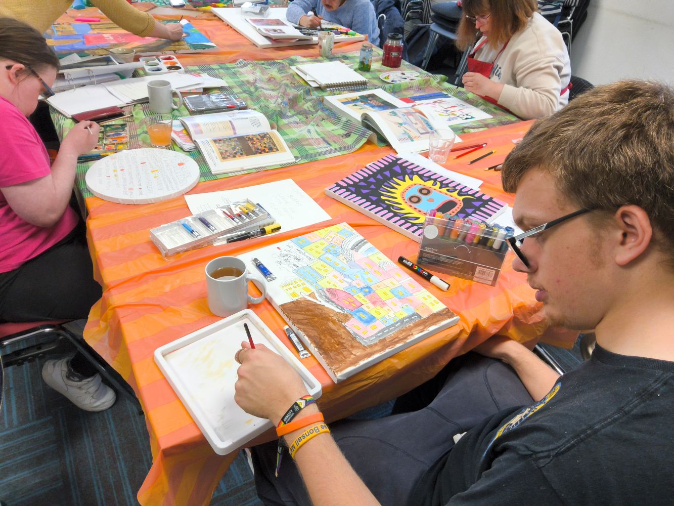 People seated around a table each intent upon their own art work. The table top is covered with pens and reference books