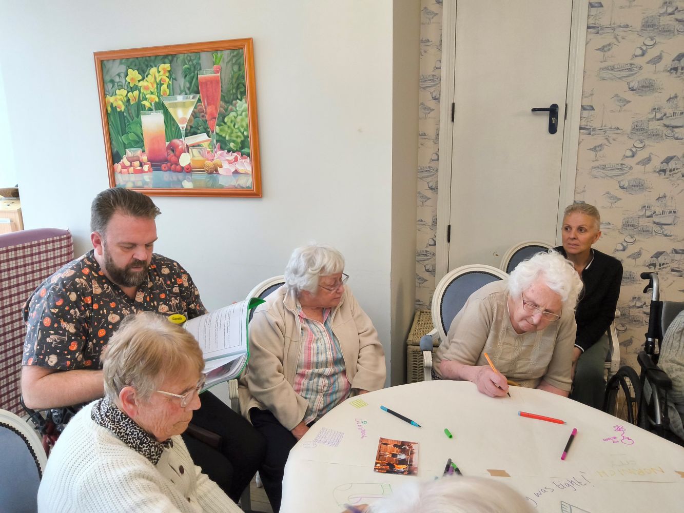 A man sits looking at a publication. On either side are women, seated at a circular table. One is drawing whilst another watches intently; the woman on his right might also be drawing. On the wall is a picture of three glasses and daffodils, and against another wall there's a narrow white door with a dark handle; a woman in a dark top sits by the wall, slightly removed from the table