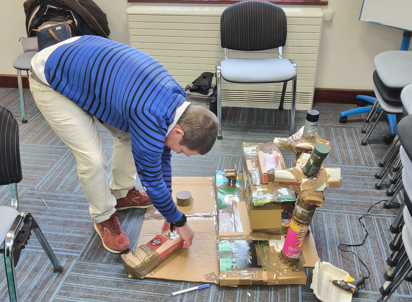 A man in blue and black striped top and light trousers bends over to stick a cardboard shape onto a model he's creating out of boxes, tubes and thick brown sticky tape