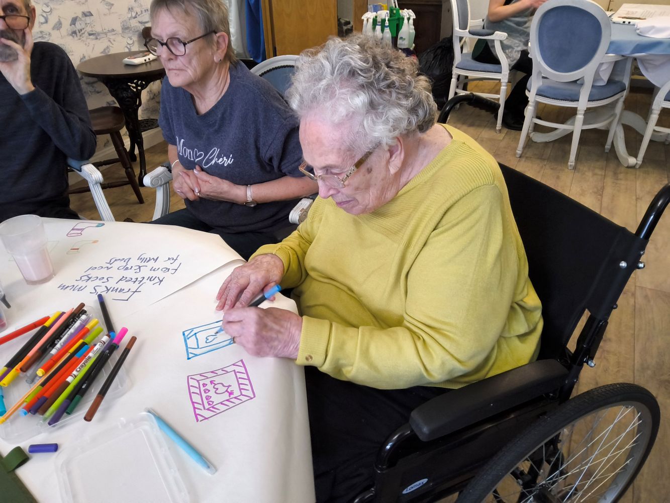 A woman seated in a dark wheel chair and wearing a mustard coloured top looks intently as she draws a turquoise picture on the paper in front of her. A woman in a blue T-shirt and wearing glasses is seated beside her. Numerous coloured felt tip pens lie on the table in front of them
