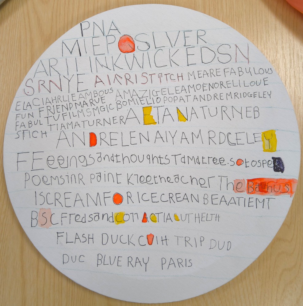 A white circular canvas covered in letters some of which spell out words others are random and some are picked out in yellow or orange paint