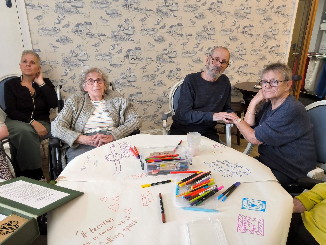In the foreground is a circular table, covered with lining paper. Numerous coloured felt tip pens lie scatted on the table and there are pictures and lines of text written in different styles. A couple sits on the righthand side, the gentleman leaning in slightly and holding hands with the woman on his left. Another woman dressed in a light coloured cardigan and top stares straight at the camera, her hands crossed in her lap. Behind and to her right sits a woman in a dark top and trousers. The wall behind them has a motif of blue boats and beach huts