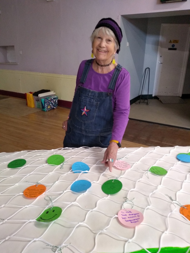 A woman wearing a purple hat, purple top and denim dungarees points to a circle of green card on which she has written a personal goal. The circle is on a table covered with a football net and there are other different coloured goals tied to it