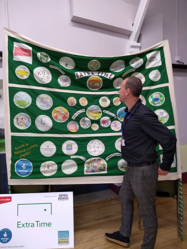 A man stands in front of a large green football banner decorated with embroidered circles, slogans and logos