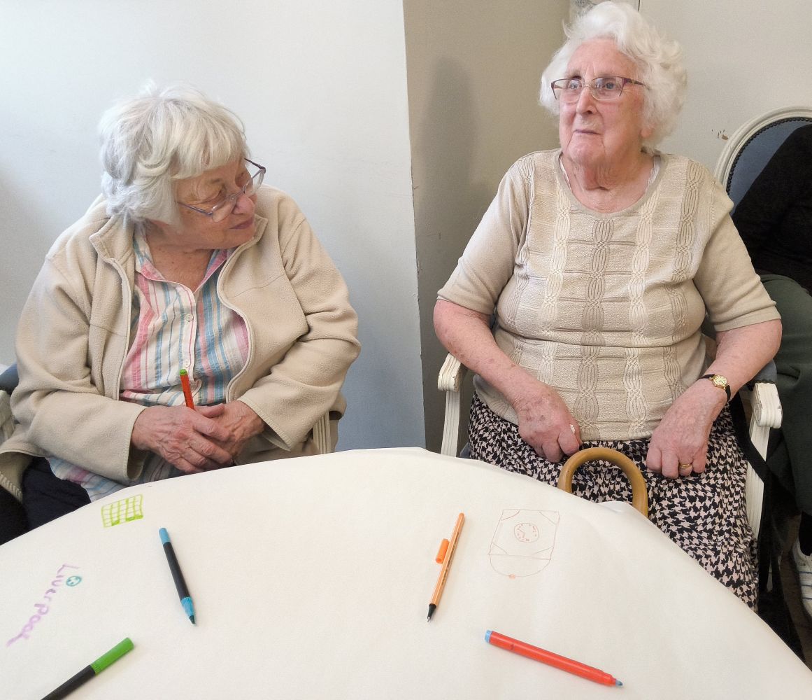 Two women sit side by side by a table on which they have been drawing images - a few pens lie scattered on the table top
