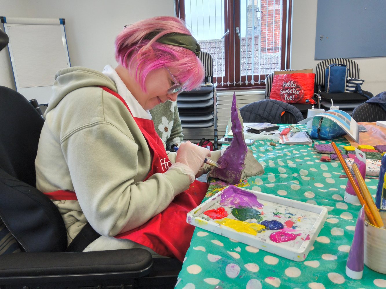 A woman with pink hair, black hairband and light hoodie paints a clay spire in purple acrylic paint