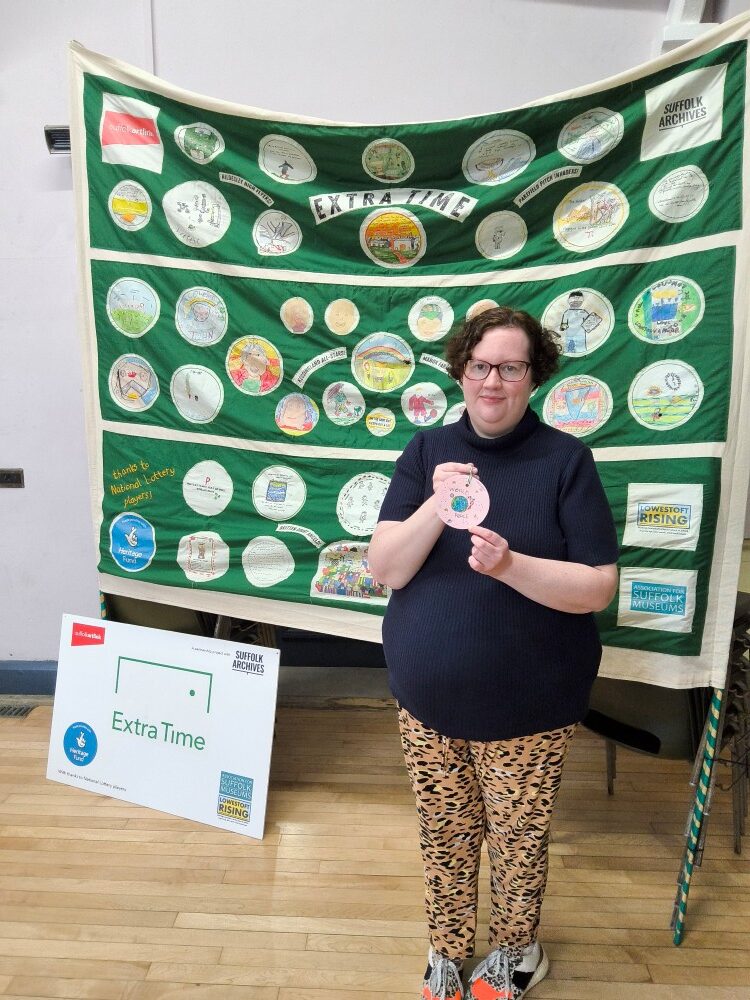 A woman stands in front of a green football banner covered with circular illustrations. She's holding a personal goal she's drawn on a pink circle of card
