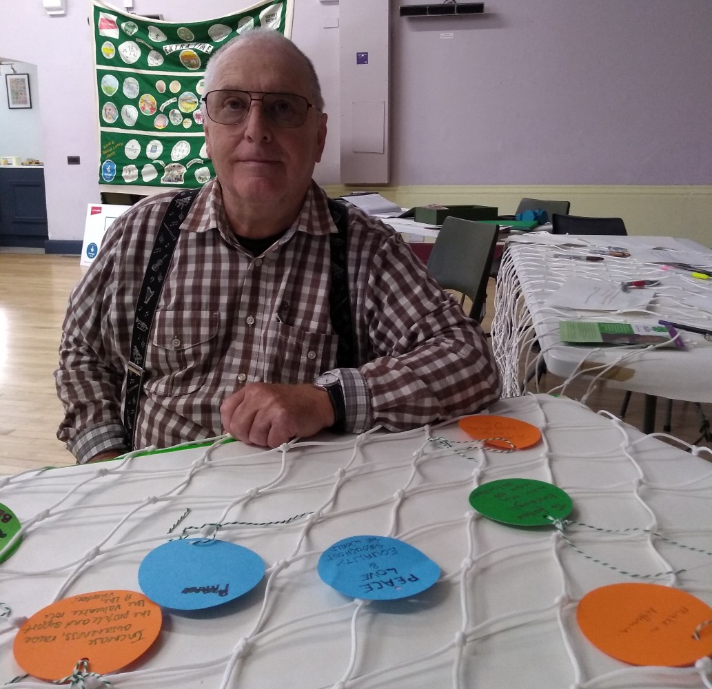 A gentleman wearing a check shirt and braces sits at a table with a piece of football net in front of him on a white background. The next has been decorated with circles of different coloured card, each bearing an individual goal