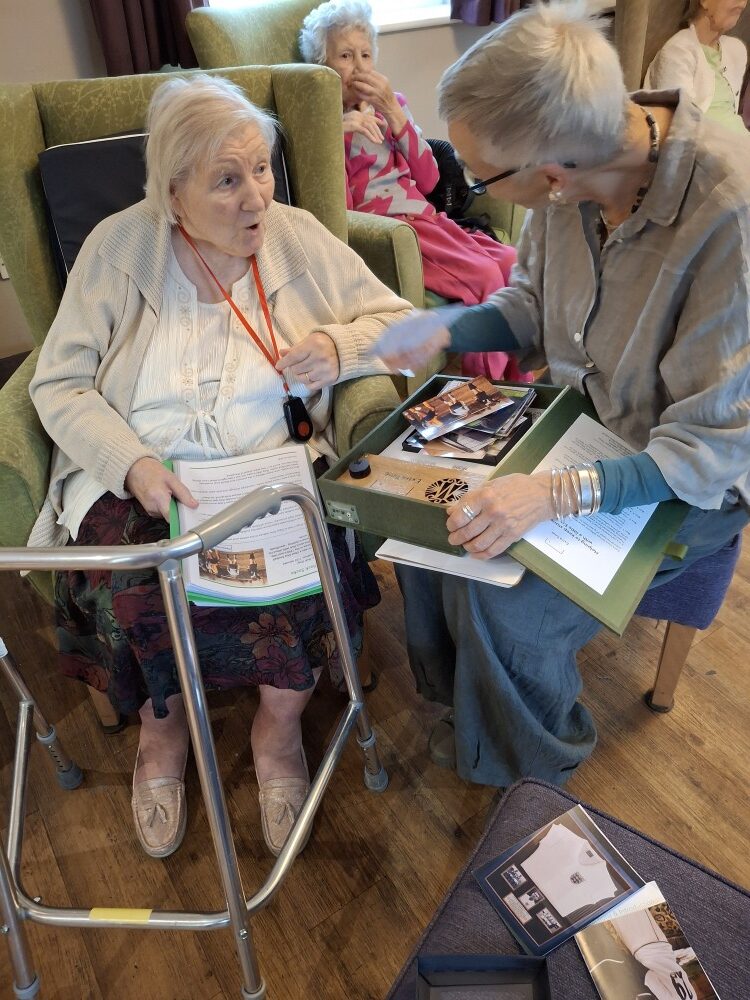 Two women sit side by side, one in a green armchair and the other on a stool. The woman in the armchair has a red lanyard around her neck and a walking frame in front of her - she is looking at the other woman with an expression of surprise on her face. The woman beside her holds a green box on her lap in which there's several photographs. In the background sits a lady dressed in pink