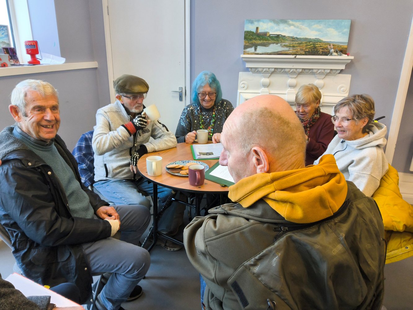 A group of adults seated around a table, drinking mugs of coffee and with a plate of biscuits. They are chatting to one another whilst a couple appear to be looking at a book. A gentleman in the right foreground, wearing a mustard coloured hoody under a green jacket is looking to the side, whilst the man seated beside him grins broadly