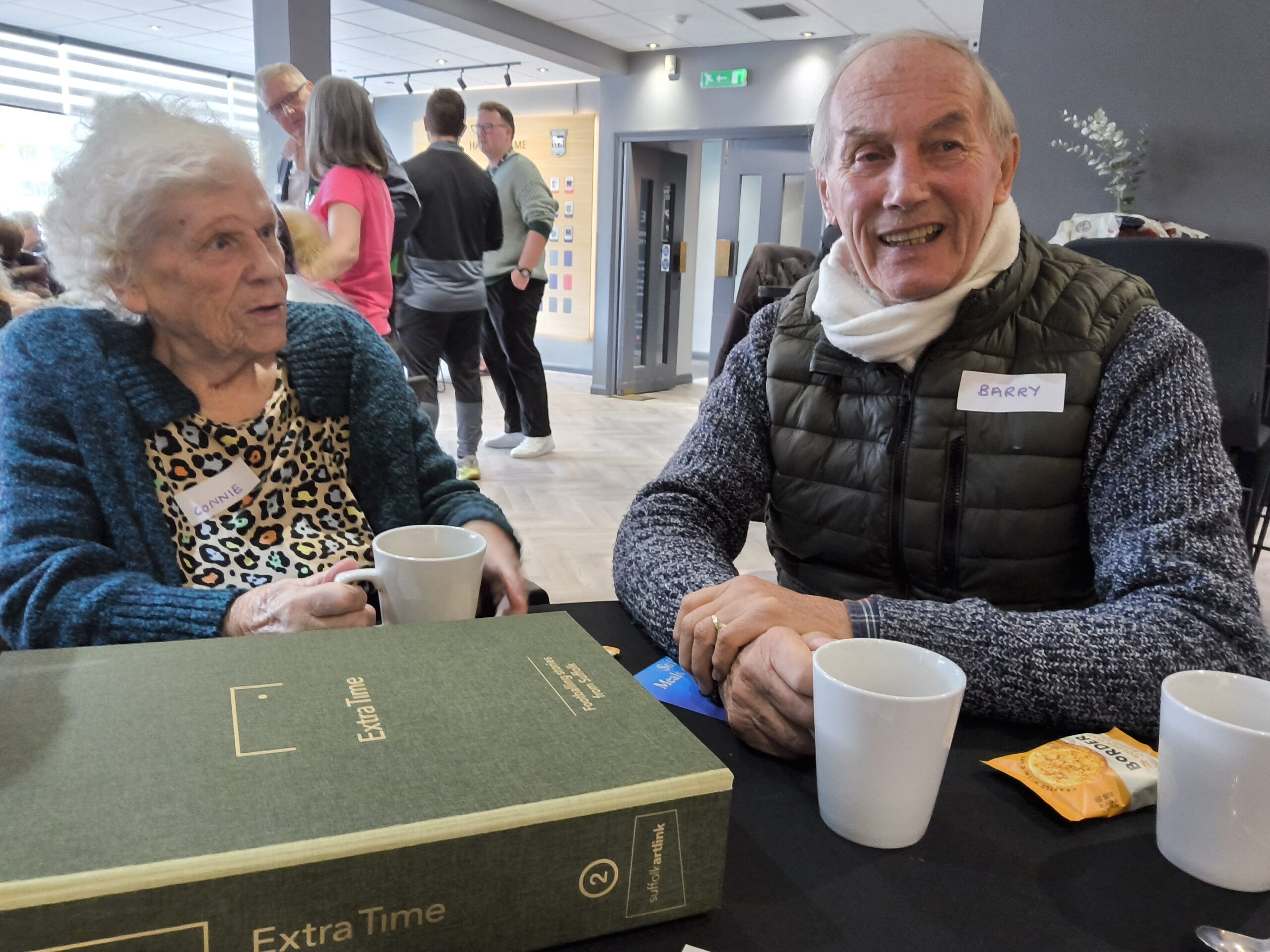 A gentleman wearing a white scarf and a black sleeveless puffa jacket over a dark jumper, smiles towards the camera, whilst a woman seated beside him looks on. In the foreground are white coffee cups and the Extra Time story box