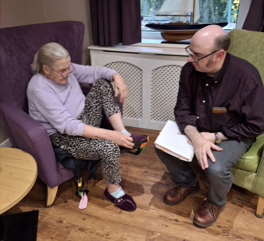 A woman sits in a purple armchair beside a man dressed in dark shirt and trousers, with a writing pad on his lap. The woman holds her foot up in the air as if she's adjusting her purple slippers