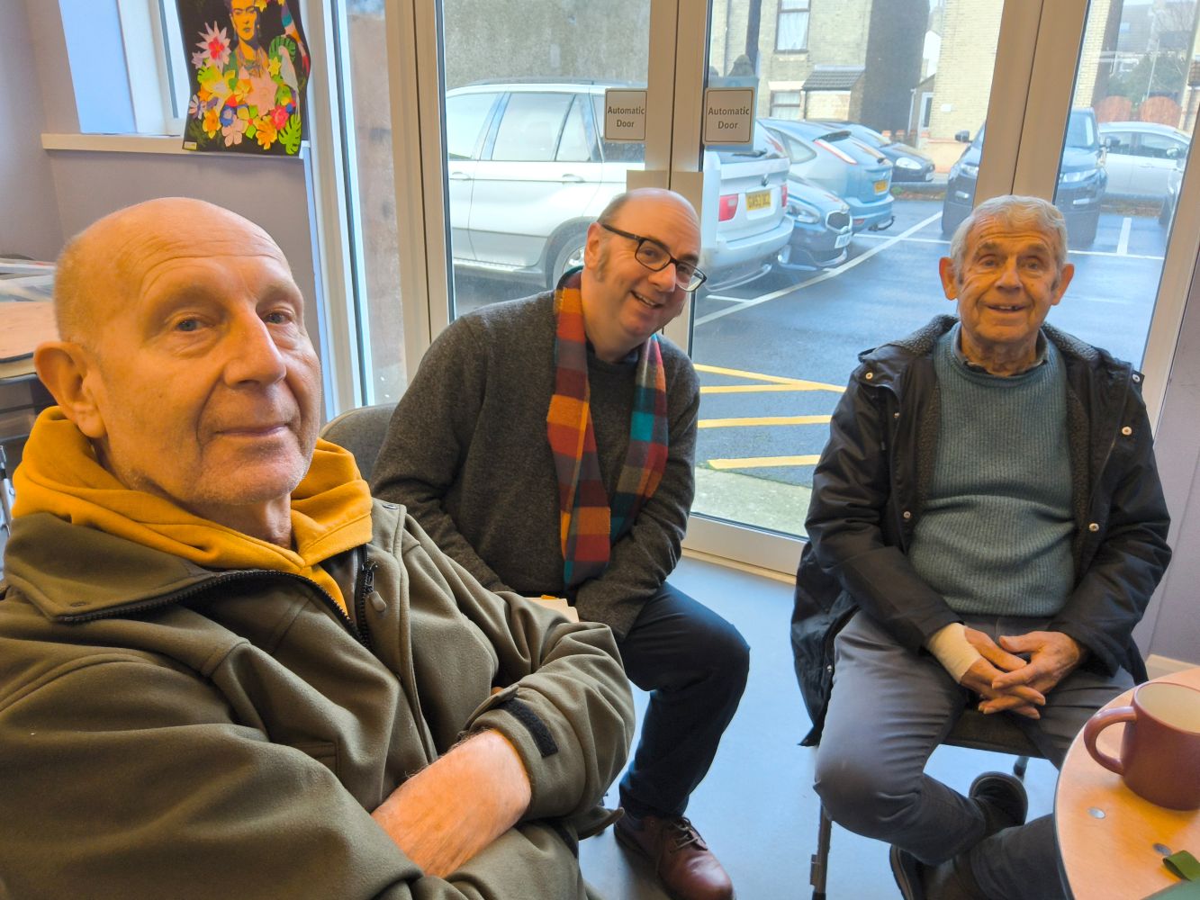 A row of three gentlemen seated, looking towards the camera. The chap in the middle is wearing a scarf of many colours. Large glass doors behind open to a view of a car park with several vehicles in place