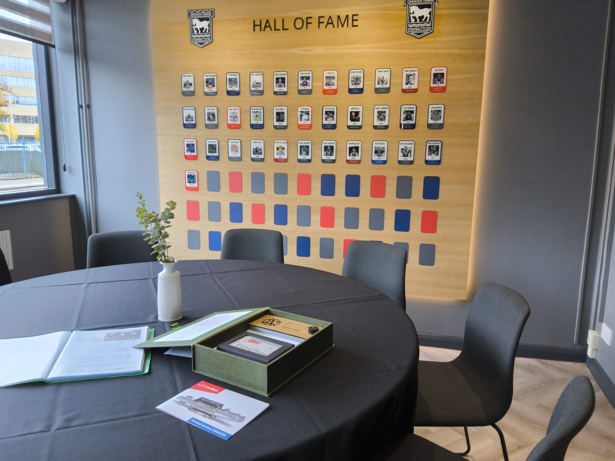 A large Ipswich Town Hall of Fame board fixed to the wall, and in the foreground a circular table covered in a dark cloth and the Extra Time Story Box beside a vase with a single spray of greenery