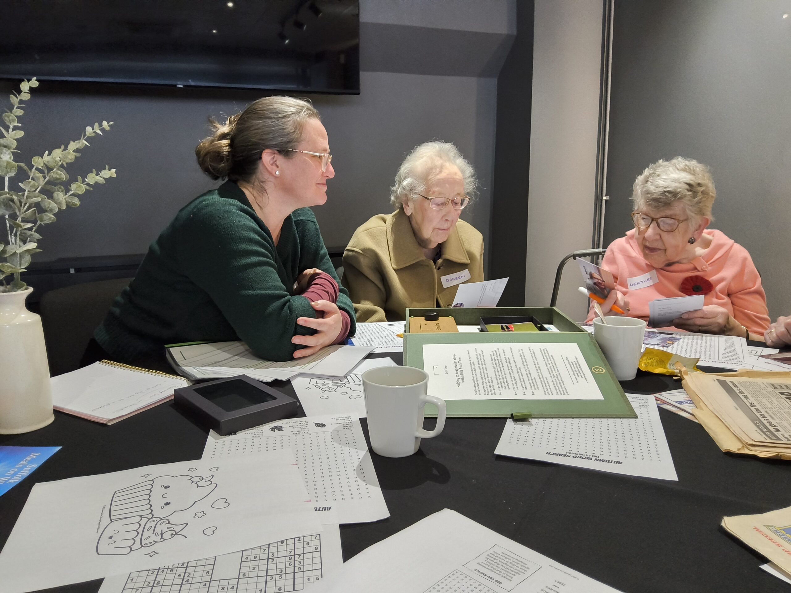 Two elderly ladies, one wearing a camel-coloured coat, are focused intently on some photographs they're holding, whilst another woman in a dark green jumper looks on. In front of them on the table is an array of papers and white coffee mugs