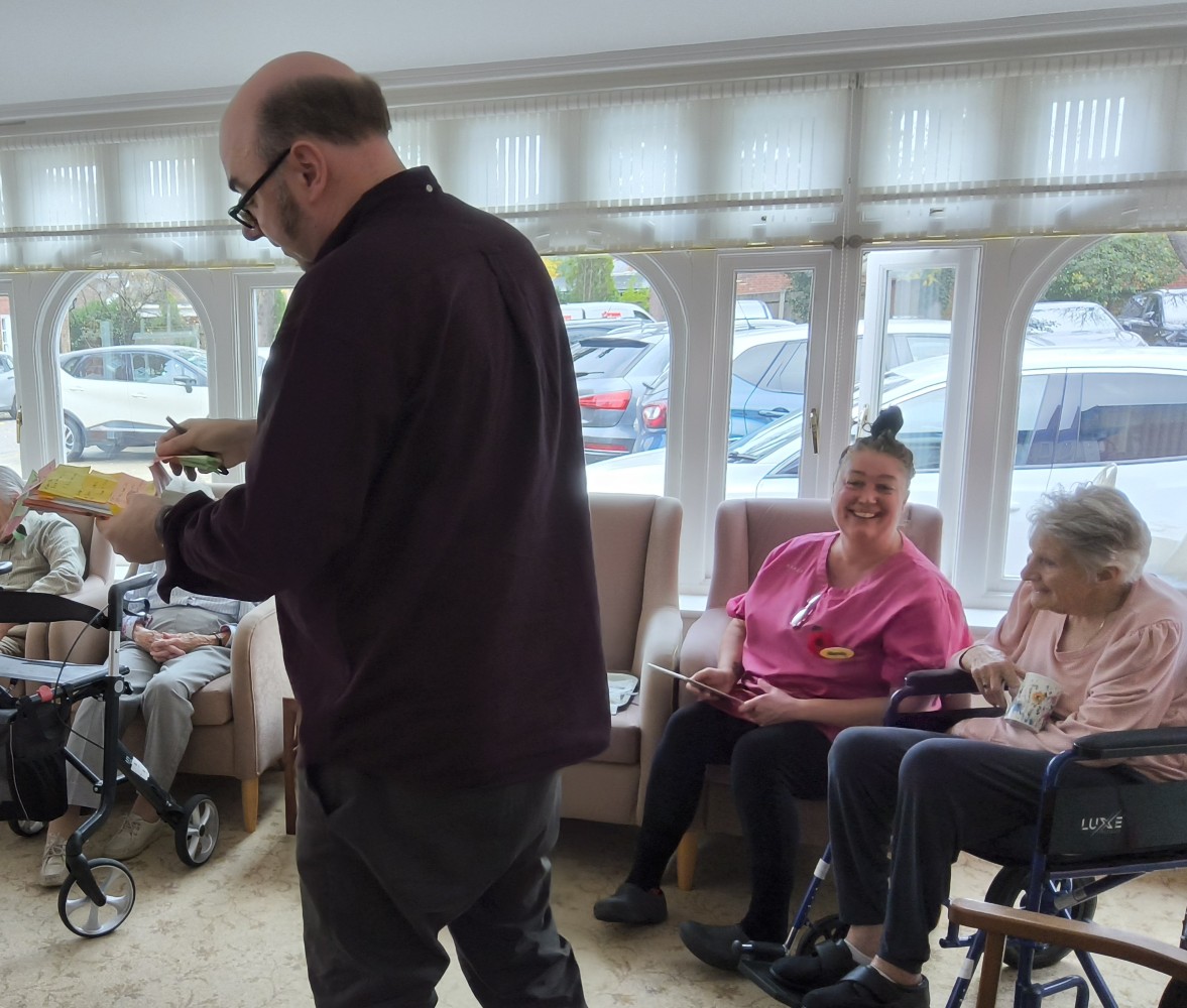 Interior scene at a residential care home. A member of staff sits next to a resident in a wheelchair, smiling at the camera. Her hair is put up in a bun on top of her head and a pair of glasses stuck in the front of her pink top reflect the light and gleam. In the foreground stands a man, dressed in dark clothing holding a book and pen in his hands