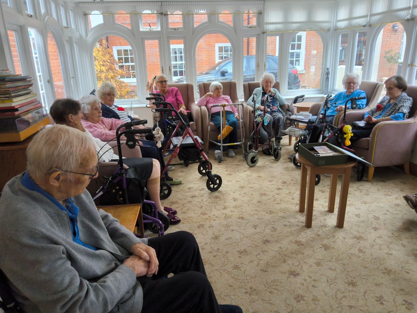 A group of residents seated a large sun lounge with tall windows, . There's a variety of walking frames dotted around and in the middle of the floor is a small circular table with an open box file on it