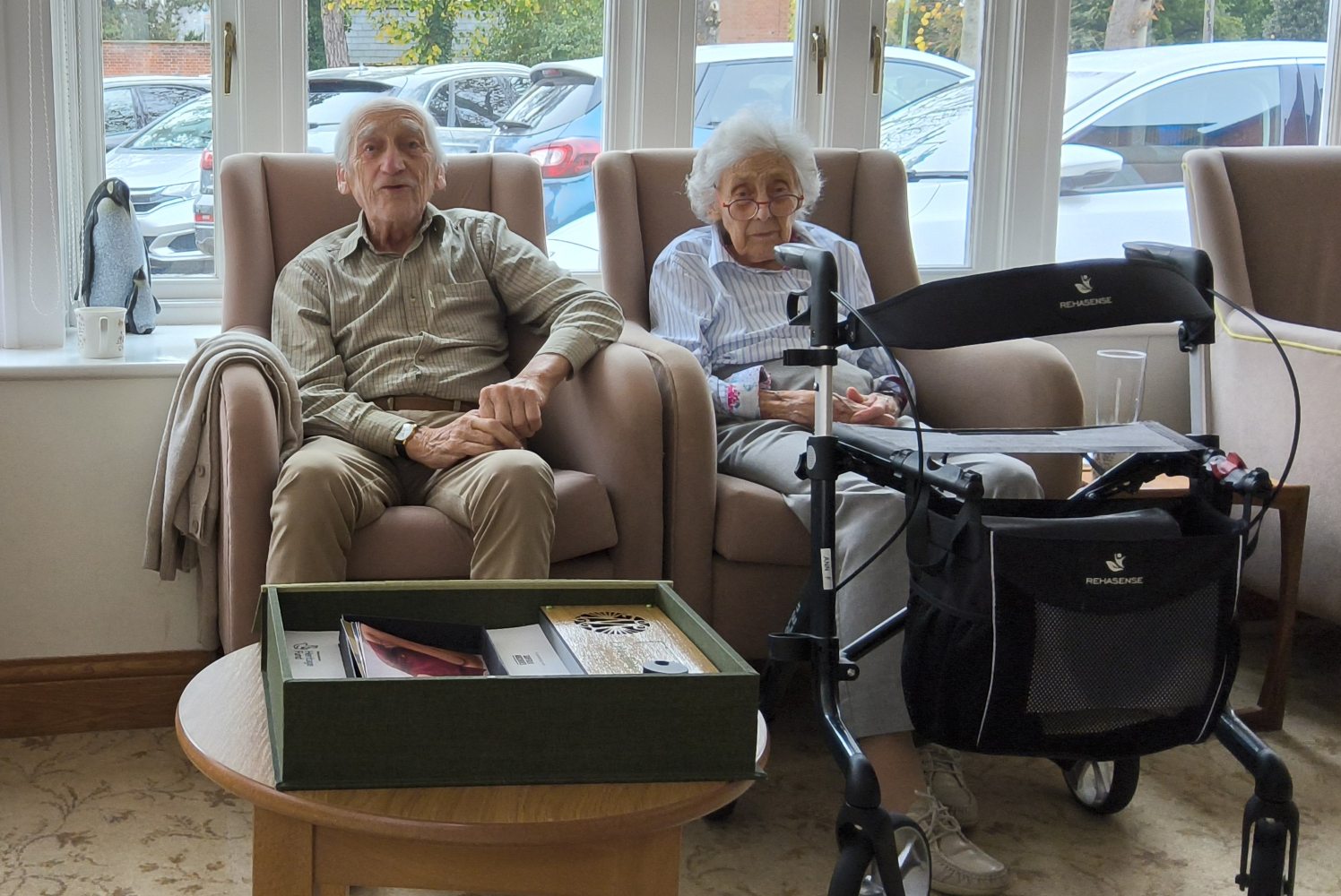 Two residents seated side by side. The gentleman looks directly at the camera and was singing a song at the time the picture was taken. To his right on the window sill is a pair of penguins