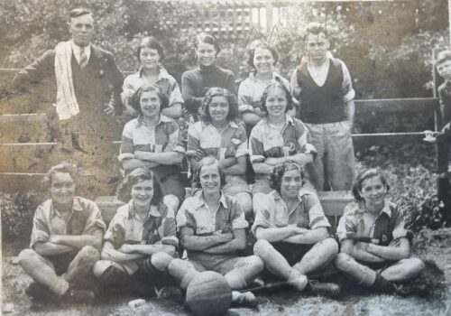 A monotone photograph of a women's football team, circa 1925, with three rows of women in football kit flanked on either side by a man, one in a dark waistcoat and the other with a towel over his shoulder