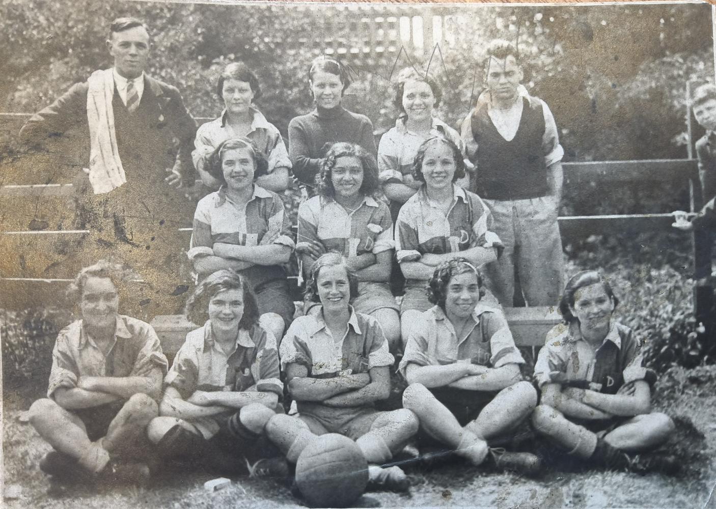 A monotone photograph of a women's football team, circa 1925, with three rows of women in football kit flanked on either side by a man, one in a dark waistcoat and the other with a towel over his shoulder